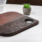 Wooden cutting board with a handle on a white surface next to a small potted plant.