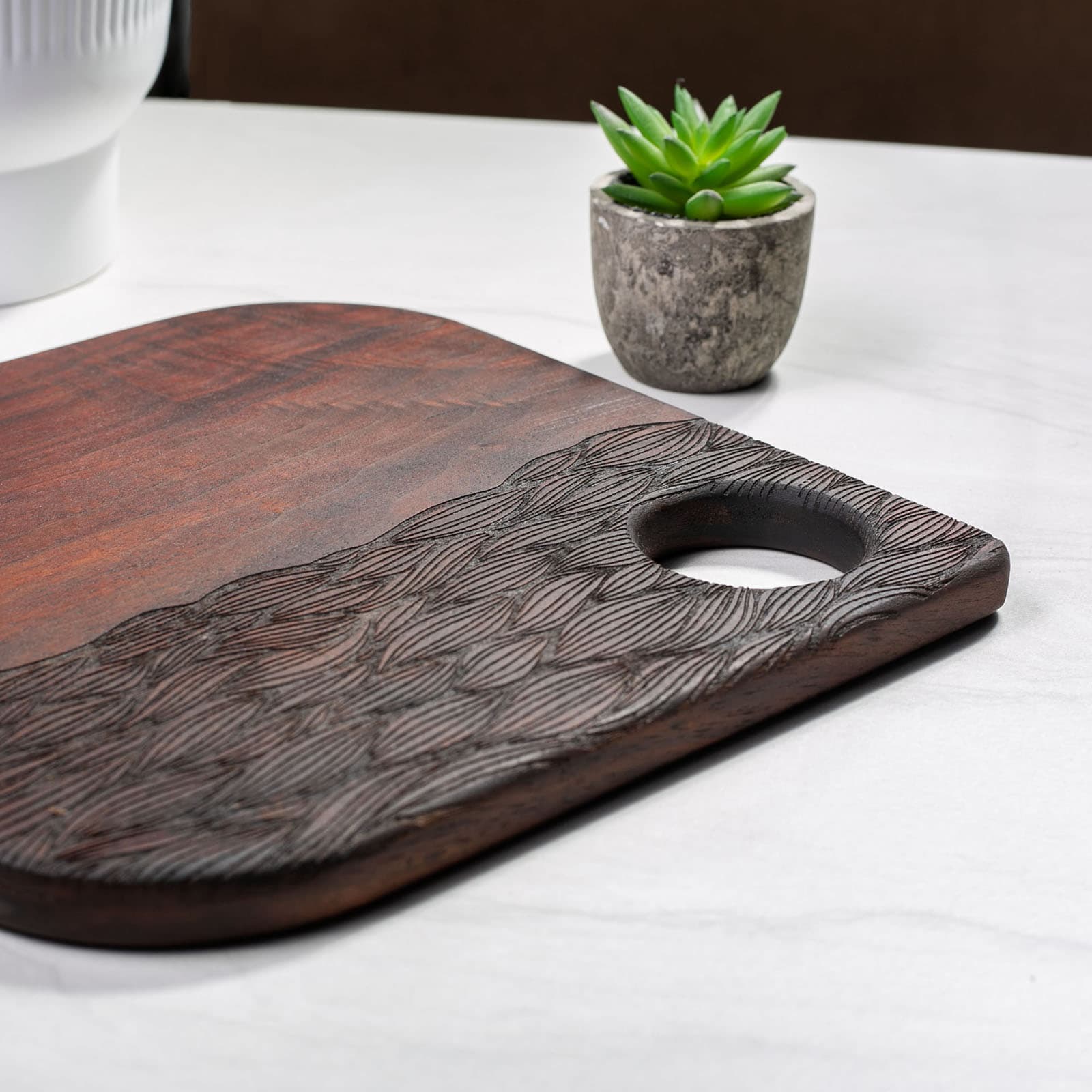 Wooden cutting board with a handle on a white surface next to a small potted plant.